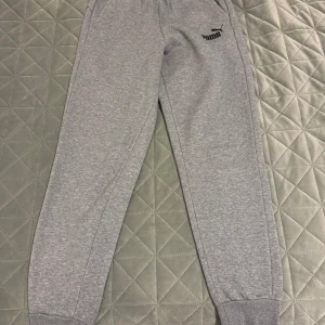 Grå mjukisbyxor från Puma - Säljer ett par grå mjukisbyxor från Puma i mycket bra skick. De är superbekväma med elastisk midja och snörning för perfekt passform. Byxorna har ribbade muddar vid bensluten och en klassisk Puma-logga på benet. Perfekta för chill eller träning!