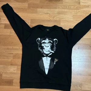 Svart sweatshirt från Mr originals - Säljer en cool svart sweatshirt från mr Originals i storlek M. Den har ett unikt tryck av en apa i kostym med en cigarr, vilket ger en rolig och edgy look. Perfekt för vardagsbruk eller en avslappnad fest. Tröjan är i bra skick och redo för nya äventyr!. Väldigt sällsynt då märket slutades säljas 2020. Tar även byten.