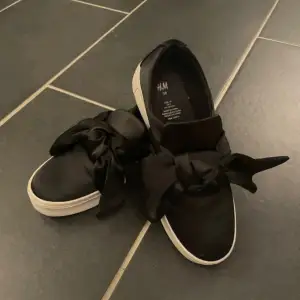 Säljer ett par svarta sneakers från H&M i bra skick. De har en snygg rosett på ovansidan och en vit sula. Perfekta för en casual look och passar till många olika outfits.