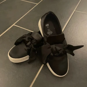 Svarta sneakers med rosett från H&M - Säljer ett par svarta sneakers från H&M i bra skick. De har en snygg rosett på ovansidan och en vit sula. Perfekta för en casual look och passar till många olika outfits.