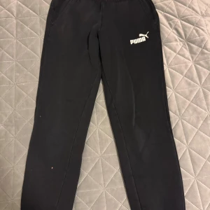 Svarta mjukisbyxor från Puma - Säljer ett par svarta mjukisbyxor från Puma i bra skick. Byxorna har en elastisk midja och ribbade muddar vid bensluten för en bekväm passform. Perfekta för träning eller en avslappnad dag hemma. Några små märken finns, men inget som stör funktionen.