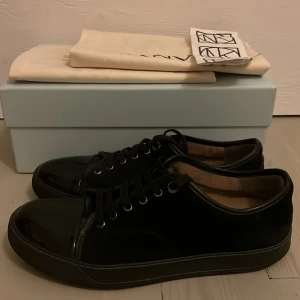 Svarta sneakers från Lanvin - Säljer ett par svarta Lanvin sneakers i lackmaterial. De är i mycket bra skick och kommer med originalkartong och dustbag. Perfekta för både vardag och fest med sin stilrena design och glansiga yta. Skorna har snörning och en bekväm passform.| Kvitto medföljer såklart ✌️👍| en liten defekt på undersidan ett hål men märks inte så mycket vid användning ✅