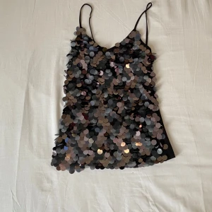 Svart paljettlinne - Säljer ett svart linne med tunna axelband och massor av glittriga paljetter på framsidan. Perfekt för festliga tillfällen när du vill sticka ut! Linnet har en tight passform och är i mycket bra skick. Passar perfekt till jeans eller kjol för en snygg partylook.