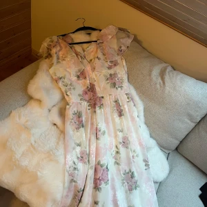 Blommig klänning med volangdetaljer - Säljer en superfin blommig klänning med volangdetaljer. Den har en v-ringad desig. Klänningen är i ljusa toner med rosa och gröna blommor på. Den är ärmlös och har en luftig passform som gör den bekväm att bära hela dagen. Använd en gång på studentbalen och alla älskade den🥹🩷💖💕💓Nyrpris är 1200 kr 