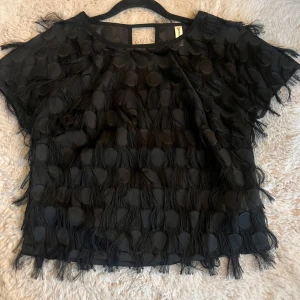 Svart topp med fransar - Snygg svart topp med korta ärmar och coola fransdetaljer. Perfekt för en festlig kväll eller när du vill sticka ut lite extra. Den har en unik textur med cirkulära mönster och fransar som ger en trendig look.