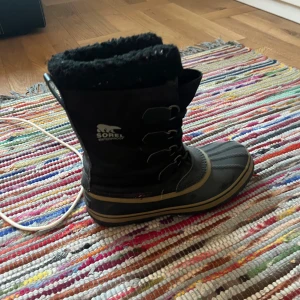 Svarta vinterstövlar från Sorel - Säljer ett par svarta Sorel vinterstövlar i bra skick. De är fodrade med mjukt material för extra värme och har snörning framtill. Perfekta för kalla vinterdagar med sin vattentäta design. Stövlarna är robusta och bekväma, idealiska för snö och slask.