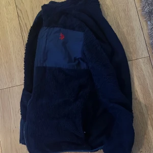 Mörkblå fleecejacka från U.S. Polo Assn. - Säljer en mysig mörkblå fleecejacka från U.S. Polo Assn. Perfekt för kyliga dagar med sin mjuka och varma känsla. Jackan har en dragkedja framtill och en liten röd logga på bröstet. Passar perfekt för höst och vår! Nypris ca 2000. Pris kan diskuteras