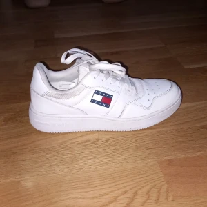 Vita sneakers från Tommy Jeans - Säljer ett par vita sneakers från Tommy Jeans i mycket bra skick. Skorna har en klassisk design med snörning och det ikoniska Tommy Jeans-märket på sidan. Perfekta för både vardag och festliga tillfällen. De är bekväma och stilrena, ett måste i garderoben!