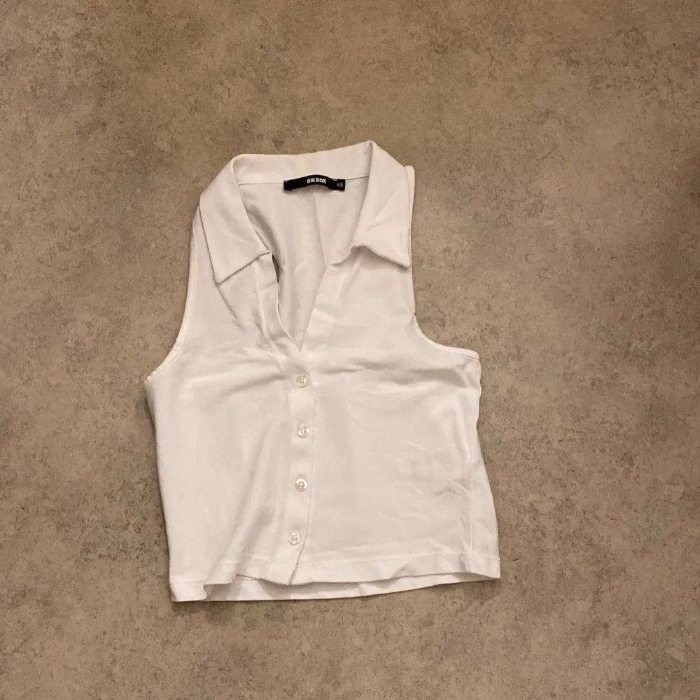 Säljer en stilren vit ärmlös topp från BikBok i storlek XS. Toppen har en klassisk krage och knappar framtill, vilket ger en snygg och enkel look. Perfekt för sommardagar eller som en del av en lager-på-lager outfit. 🕶️. Yläosat.