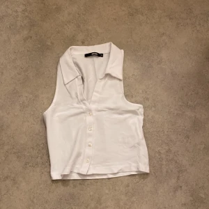 Vit ärmlös topp från BikBok - Säljer en stilren vit ärmlös topp från BikBok i storlek XS. Toppen har en klassisk krage och knappar framtill, vilket ger en snygg och enkel look. Perfekt för sommardagar eller som en del av en lager-på-lager outfit. 🕶️