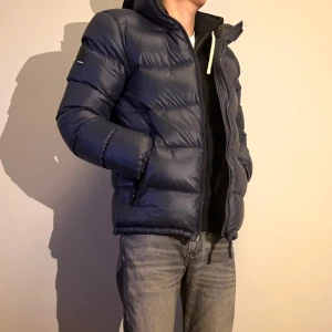 Blå dunjacka från Superdry - Säljer en svart dunjacka från Superdry i mycket bra skick. Jackan är perfekt för kalla dagar med sitt dunfoder och har en dragkedja framtill. Den har en normal passform, och är varm vilket gör den bra inför vintern! 