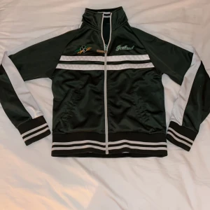 Grön och vit jacka med Irland-broderi - Säljer en grön och vit zip up med broderad text 'Irland' på bröstet och ryggen. Jackan har dragkedja framtill och långa ärmar med vita detaljer.!