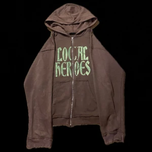 Brun hoodie med tryck från Local Heroes - Säljer en brun hoodie från Local Heroes med coolt grönt tryck på framsidan. Den har en dragkedja och en skön luva, perfekt för chilliga dagar. Passar bra till både jeans och joggers. Hör av dig om du är intresserad! 💚