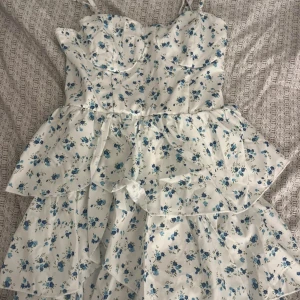 Vit klänning med blå blommor - En super fin klänning med blåa blommor på. Rosetter på banden som man kan sjustera. Aldrig använd! 