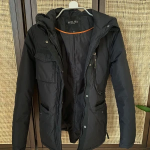 Svart vinterjacka från Hollies - Säljer en svart vinterjacka från Hollies i mycket bra skick. Jackan har en praktisk huva och flera fickor med dragkedja och knappar. Perfekt för kalla vinterdagar. Den är långärmad och har en snygg, figurnära passform. Passar både till vardags och lite finare tillfällen.