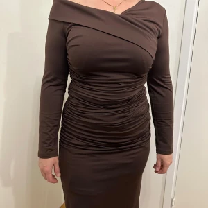 Brun långärmad klänning - Snygg brun klänning med lång ärm och offshoulder-design. Den har en tight passform med rynkade detaljer som ger en smickrande siluett. Perfekt för höst- och vinterfester!
