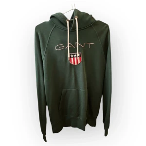 Grön hoodie från GANT - Säljer en snygg grön hoodie från GANT i storlek M. Den har en klassisk design med GANT-loggan och ett coolt emblem på framsidan. Perfekt för höst och vår med sin mjuka och bekväma passform. Huvan har vita snören och en känguruficka framtill. Passar både till vardags och chill.