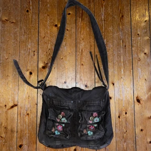 Brun axelväska med broderade blommor - Säljer en snygg brun axelväska med coola broderade blommor på fickorna. Från Åhléns.  Väskan har två stora fickor framtill med knappar och en justerbar axelrem. Insidan är fodrad i brunt tyg med en dragkedja för extra säkerhet. Har även en ficka på insidan. Köparen står för frakt, skickar spårtbart PostNord paket.