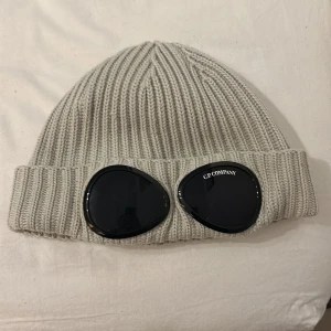 Grå ribbad mössa från C.P. Company - Snygg grå ribbad mössa från C.P. Company med coola svarta glasögondetaljer. Perfekt för att hålla sig varm och stilren under kyliga dagar. Mössan är i ett mjukt och bekvämt material, idealisk för både vardag och fest. Säljs på grund av att jag helt enkelt har tröttnat på den! Tveka inte på att höra av dig om du har några frågor!