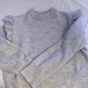 Grå tröja med volangdetaljer från Gina Tricot - Superfin grå tröja från Gina Tricot i storlek 146/152. Den är stickad och har långa ärmar med volangdetaljer vid axlarna. Perfekt för höst och vinter när du vill vara både varm och stilren. Passar till både vardag och festliga tillfällen!