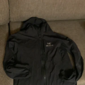 Svart vindjacka från Arc'teryx - Säljer en svart vindjacka från Arc'teryx i bra skick. Jackan har en praktisk huva och dragkedja framtill. Perfekt för blåsiga dagar och utomhusaktiviteter. Passar både till vardags och träning. Har även ett litet hål vid tricep muskeln