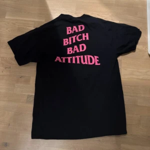 Svart t-shirt med rosa text - Cool svart t-shirt från Fashion Nova med texten 'BAD BITCH BAD ATTITUDE' i rosa på baksidan. Perfekt för en avslappnad stil med attityd. T-shirten är kortärmad och i bomull, vilket gör den bekväm att bära hela dagen.