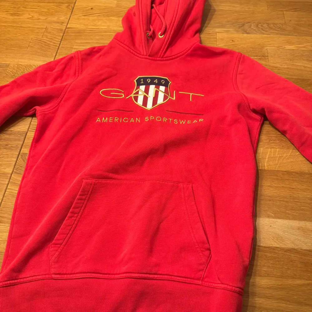 Säljer en snygg röd hoodie från GANT med tryck framtill. Den har en praktisk magficka och justerbar huva med snören. Perfekt för en avslappnad stil.. Hupparit & Collegepaidat.