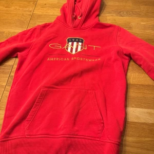 Röd hoodie från GANT - Säljer en snygg röd hoodie från GANT med tryck framtill. Den har en praktisk magficka och justerbar huva med snören. Perfekt för en avslappnad stil.