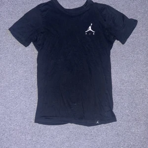 Svart t-shirt från Jordan - Säljer en svart t-shirt från Jordan med det ikoniska Jumpman-logotypen på bröstet. T-shirten är kortärmad och tillverkad i ett mjukt material som ger en skön passform. Det finns ett litet hål i framsidan av tröjan som man ser på bilden. Perfekt för en sportig look eller avslappnad stil.