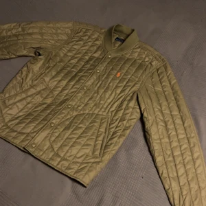 Olivgrön quiltad jacka från Polo Ralph Lauren - Säljer en snygg olivgrön quiltad jacka från Polo Ralph Lauren. Jackan har långa ärmar och en broderad logga på bröstet. Perfekt för höst och vår med sin stilrena design och praktiska knappar framtill.