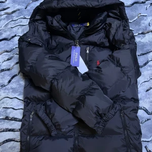 Svart dunjacka från Polo Ralph Lauren - Snygg svart dunjacka från Polo Ralph Lauren med broderad röd logotyp på bröstet. Jackan har dragkedja och praktiska fickor. Perfekt för kyliga dagar med sin varma och stilrena design.