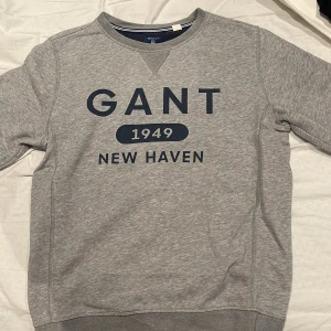 Grå sweatshirt från GANT - Säljer en stilren grå sweatshirt från GANT med tryck 'GANT 1949 NEW HAVEN' i mörkblått på framsidan. Tröjan har rund hals och långa ärmar, perfekt för en avslappnad stil.