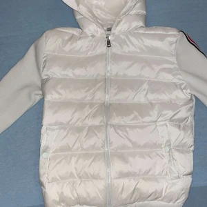 Vit Moncler cardigan - Snygg vit dunjacka från Moncler med huva och dragkedja. Jackan har långa ärmar med röd och svart randdetalj samt Moncler-logga på ärmen. Perfekt för kyligare dagar. Jackan är används några gånger inte mer än 5 gånger, priset diskuteras