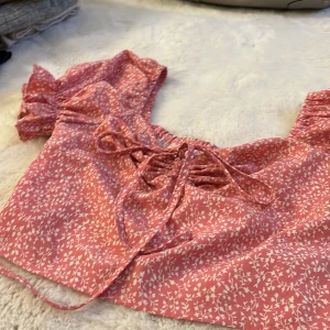 Rosa blommig topp med knytning - Charmig rosa topp med vita blommor och knytning framtill. Toppen har korta ärmar med volangdetaljer och en söt, feminin stil. Perfekt för varma sommardagar.🩷