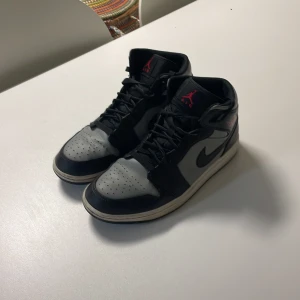 Nike Air Jordan 1 svart, grå och röd - Snygga Nike Air Jordans i svart och grått med röda detaljer. Skorna har en klassisk design med snörning och en hög siluett. Perfekta för dig som vill ha en stilren look med en sportig touch. Är även i väldigt bra skick!