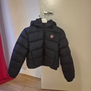 Svart vadderad jacka  från Gant - Snygg svart vadderad från Gant med dragkedja och huva. Jackan har ett stilrent märke på bröstet och är perfekt för kyligare dagar. Den är både varm och trendig, vilket gör den till ett måste i garderoben. https://www.kids-world.com/sv-se/gant-vadderad-jacka-retro-shield-svart-p-229293.html