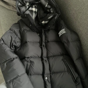 Burberry jacka  - Säljer en snygg svart dunjacka från Burberry med avtagbar huva och klassiskt rutigt foder. Jackan har dragkedja och knappar framtill samt två praktiska fickor med dragkedja. Perfekt för kyliga dagar!