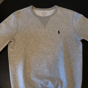 Grå sweatshirt från Polo Ralph Lauren - Säljer en stilren grå sweatshirt från Polo Ralph Lauren med rund halsringning och det klassiska logotypbroderiet på bröstet. Tröjan har långa ärmar och ribbade muddar vid ärmslut och nederkant. Perfekt för en avslappnad look.