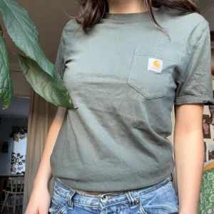 Grön Carhartt T-shirt - Säljer en grön t-shirt från Carhartt med bröstficka och logga. Perfekt för en avslappnad stil. Passar bra till jeans eller shorts. Storlek S.