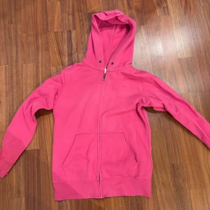 Rosa hoodie med dragkedja - Säljer en snygg rosa hoodie med dragkedja och huva. Perfekt för en avslappnad stil. Den har långa ärmar och praktiska fickor framtill. Liten i storleken. 