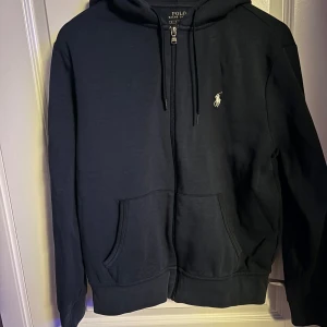 Mörkblå hoodie från Polo Ralph Lauren - Snygg mörkblå  hoodie från Polo Ralph Lauren med dragkedja och klassisk logga på bröstet. Perfekt för en avslappnad stil. Den har en huva och praktiska fickor framtill.