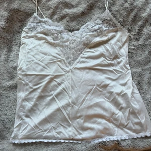 Vit satinlinne med spetsdetaljer - Snyggt vit satinlinne med tunna axelband och vacker spetsdetalj vid halsringningen. Perfekt som pyjamas linne eller ha under en kofta! 