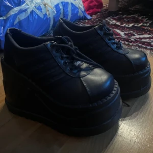 Demonia platform skor - Snygga svarta platåskor med snörning och hög sula. Perfekta för att ge lite extra höjd och stil. Skorna har en robust design med detaljerade sömmar och en klassisk look.