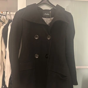 Svart kappa från Twist & Tango - Elegant svart kappa från Twist & Tango i storlek XS. Den har en klassisk design med stora knappar framtill och en stilren krage. Perfekt för kyligare dagar. Lite sprucken inuti men det går att sy.