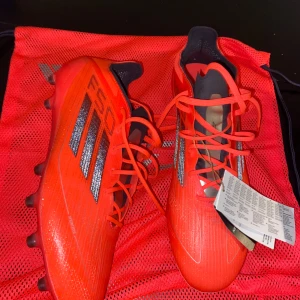 Röda fotbollsskor från Adidas - Snygga röda fotbollsskor från Adidas med svarta detaljer och snörning. Perfekta för fotbollsplanen med sina dobbar för bra grepp. Skorna kommer med en matchande röd förvaringspåse.