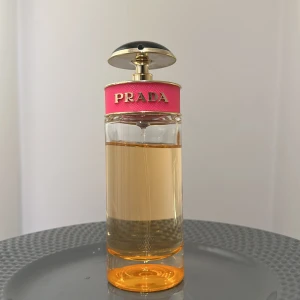 Prada Parfym Candy  - Elegant parfymflaska från Prada med en stilren design. Flaskan är genomskinlig med en gyllene vätska och har en rosa detalj med Pradas logotyp i guld. Korken är rund och glänsande, vilket ger en lyxig känsla.