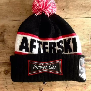 Svart mössa med texten 'Afterski' - Snygg svart mössa med texten 'Afterski' i vitt och rött. Mössan har en stor röd och vit tofs på toppen och en uppvikt kant med texten 'Bucket List'. Perfekt för kalla dagar i backen! Har aldrig använt mössan och den finns ej att köpa på internet så passa på!(frakt betalas själv)