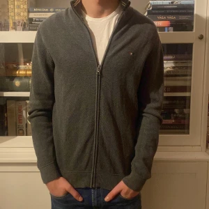 Grå kofta från Bison - Säljer en stilren grå kofta från Bison med dragkedja framtill. Koftan har en diskret logga på bröstet och är perfekt för en avslappnad look. Den är långärmad och har ribbade muddar vid ärmslut och nederkant.