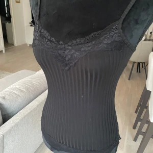 Svart ribbad linnetopp med spetsdetaljer - Snygg svart ribbad linnetopp med spetsdetaljer vid halsringningen. Perfekt för lager-på-lager eller som den är. Smala axelband och en figurnära passform.