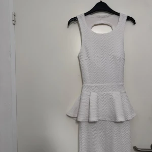 Vit ärmlös klänning med volangdetalj - Snygg vit ärmlös klänning från NLY One med volangdetalj i midjan och öppen rygg. Klänningen har en strukturerad yta och en figurnära passform som framhäver silhuetten. Perfekt för en stilren look.